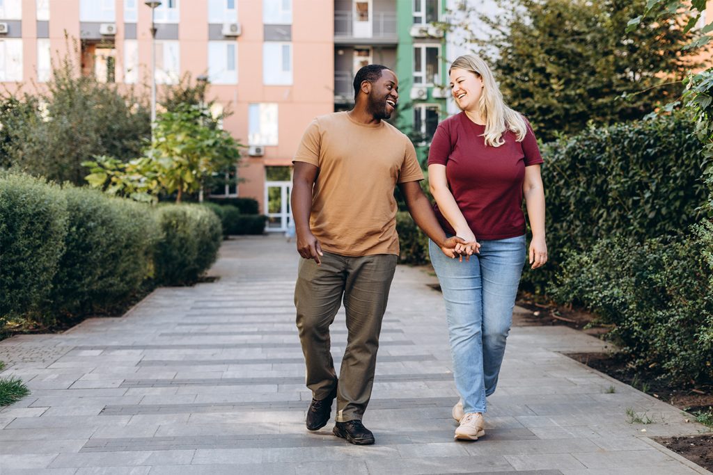 Pareja formada por un hombre negro y una mujer caucásica, cogidos de la mano y sonriendo mientras caminan por la acera frente a unos apartamentos.