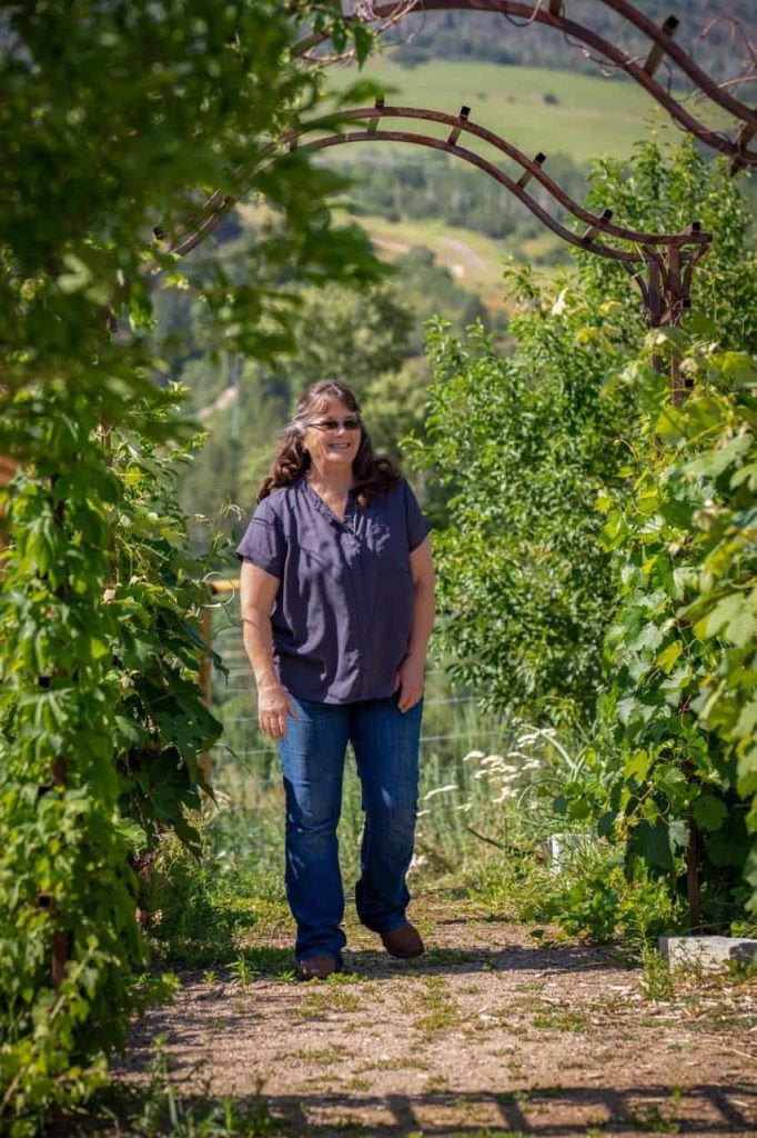 Woman smiling, walking through lush green vineyard under sunny archway.