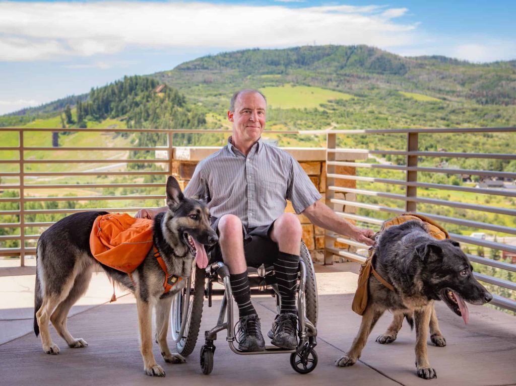 Man in wheelchair with two German Shepherds, outdoor mountain view background.