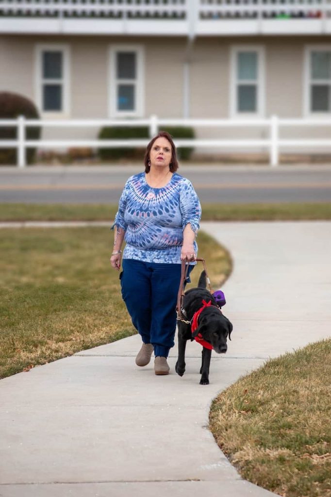 Mujer caminando con un perro guía por una acera, con un entorno suburbano al fondo.