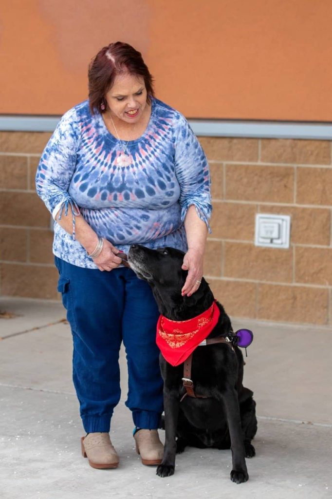 Mujer con un perro negro que lleva un pañuelo rojo, de pie frente a una pared de ladrillos.