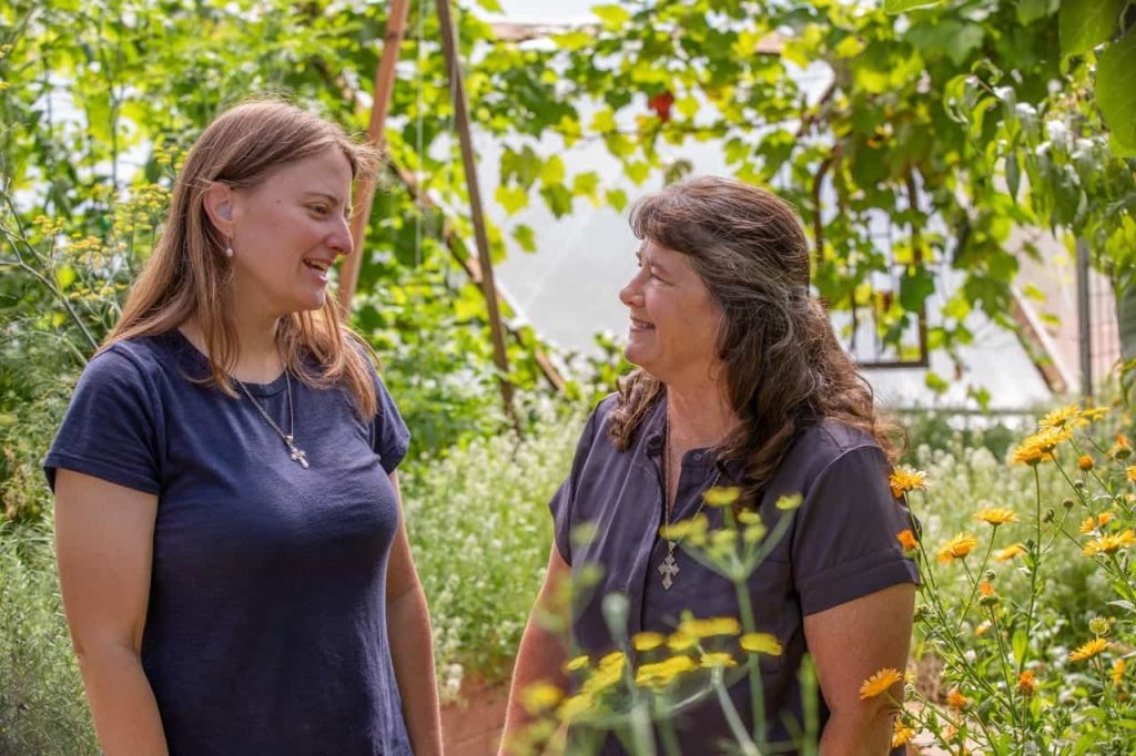 Two women smiling and talking in a lush garden, surrounded by green plants and yellow flowers.