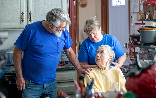 Elderly woman receiving support from caring individuals in a kitchen setting, wearing blue shirts and glasses.