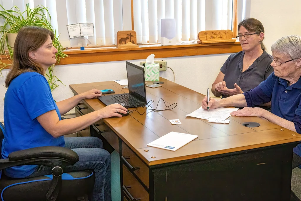 Three people having a discussion at a desk with a laptop and paperwork in an office setting.
