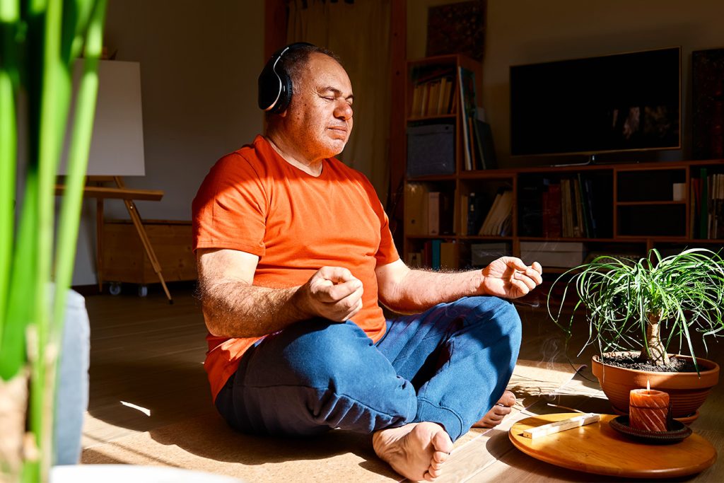 Man meditating in sunlight with headphones, surrounded by plants and incense for relaxation.