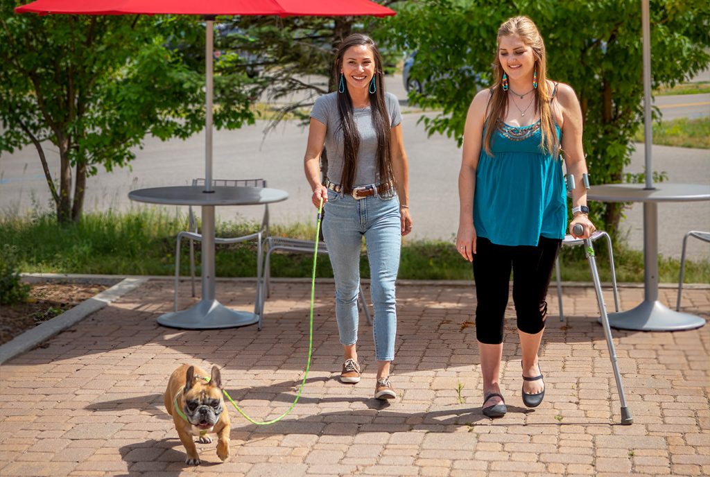 Two women walk a French Bulldog outside, one using crutches, under red umbrellas and near green trees.