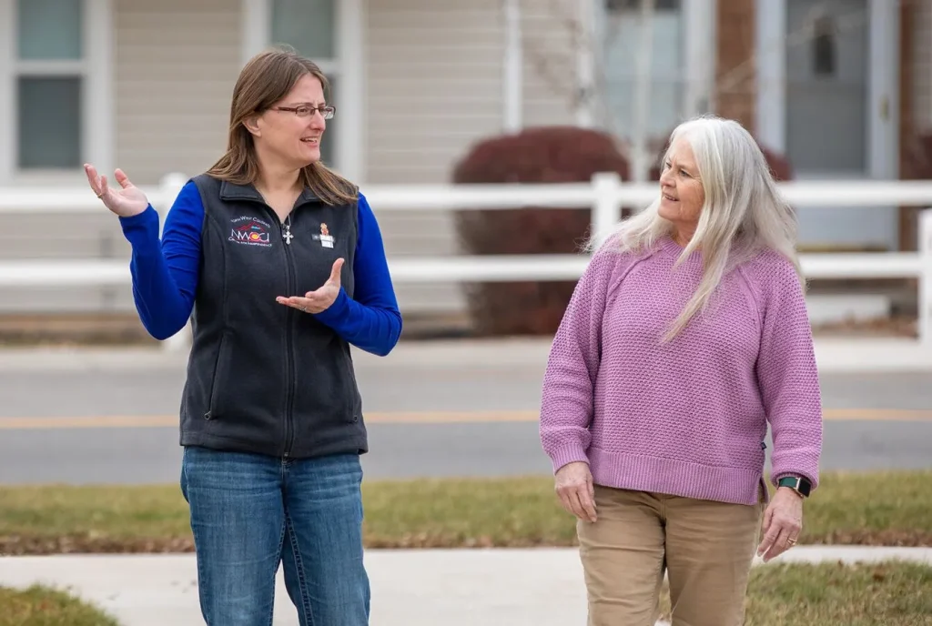 Two women having a conversation outside, one gesturing while the other listens attentively in a suburban neighborhood.