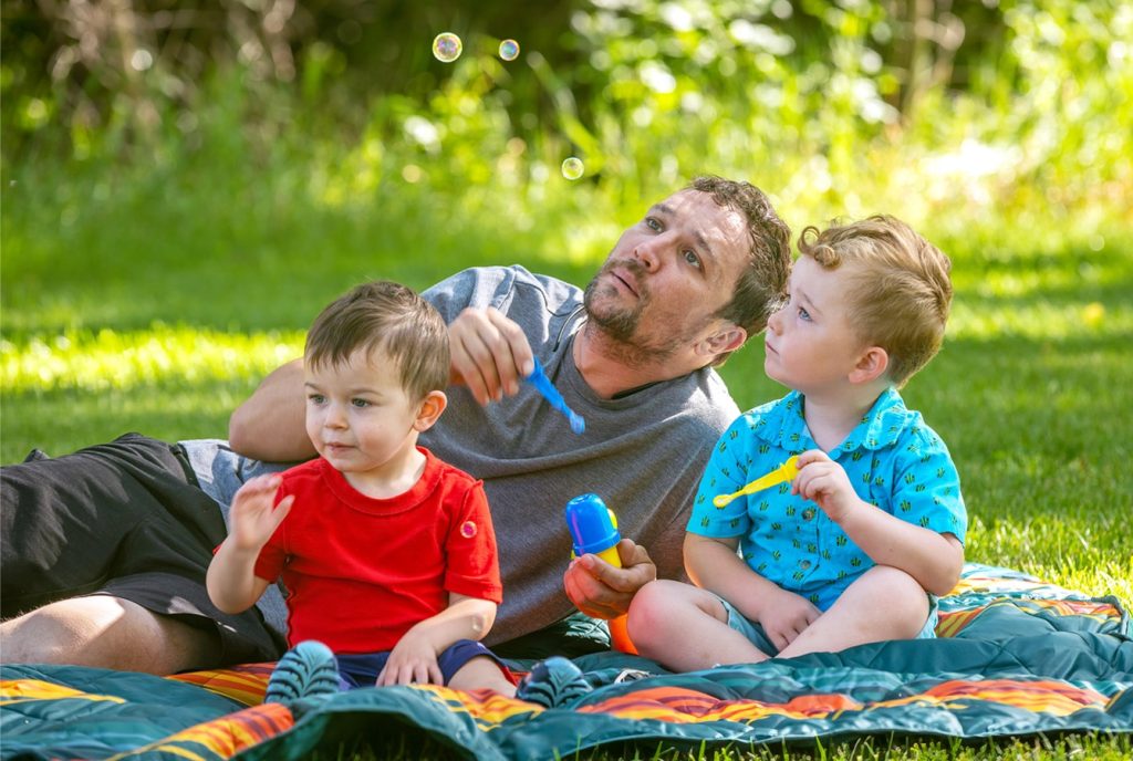 Father and sons enjoying outdoor bubble play on a sunny day picnic blanket in the park.