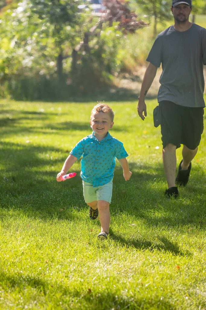 Child happily running with bubbles in sunny park, followed by adult.
