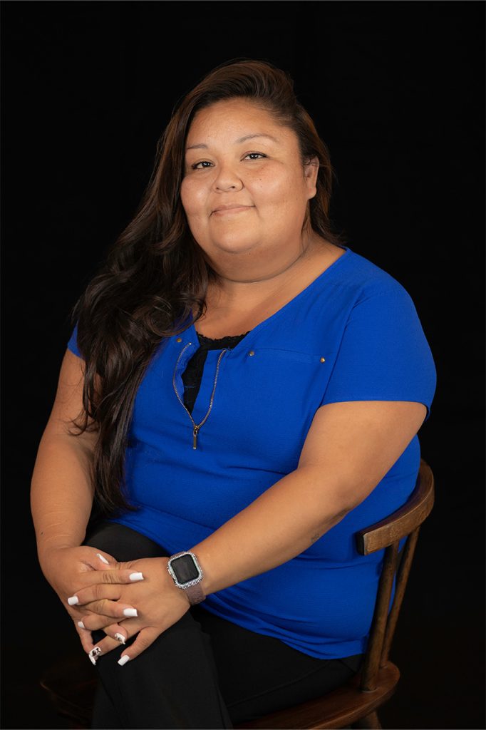 Smiling Native American woman in a blue shirt sitting on a chair against a black background.
