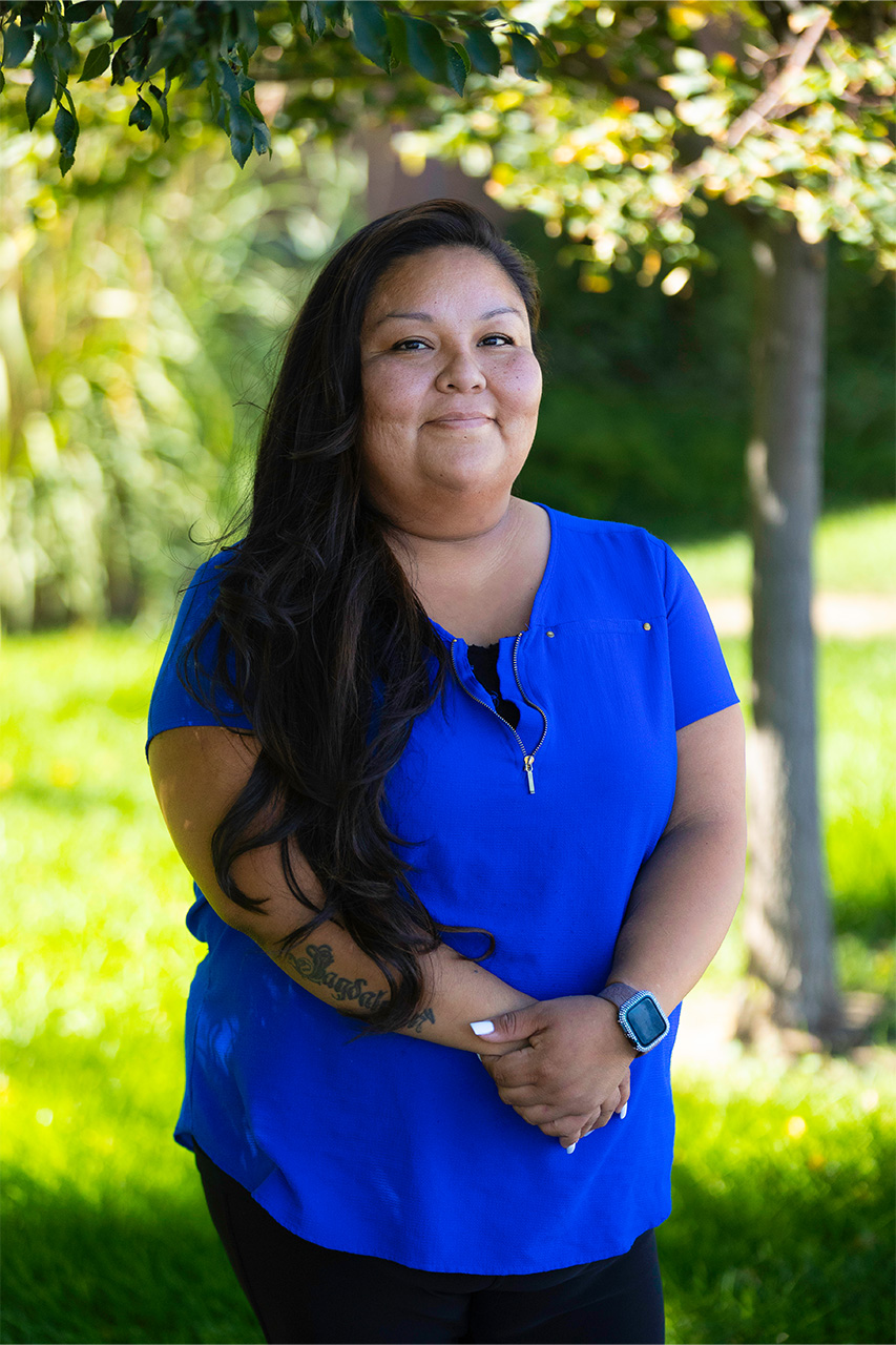 Smiling Native American woman in a blue blouse standing outdoors in a park-like setting, with sunlit greenery in the background.