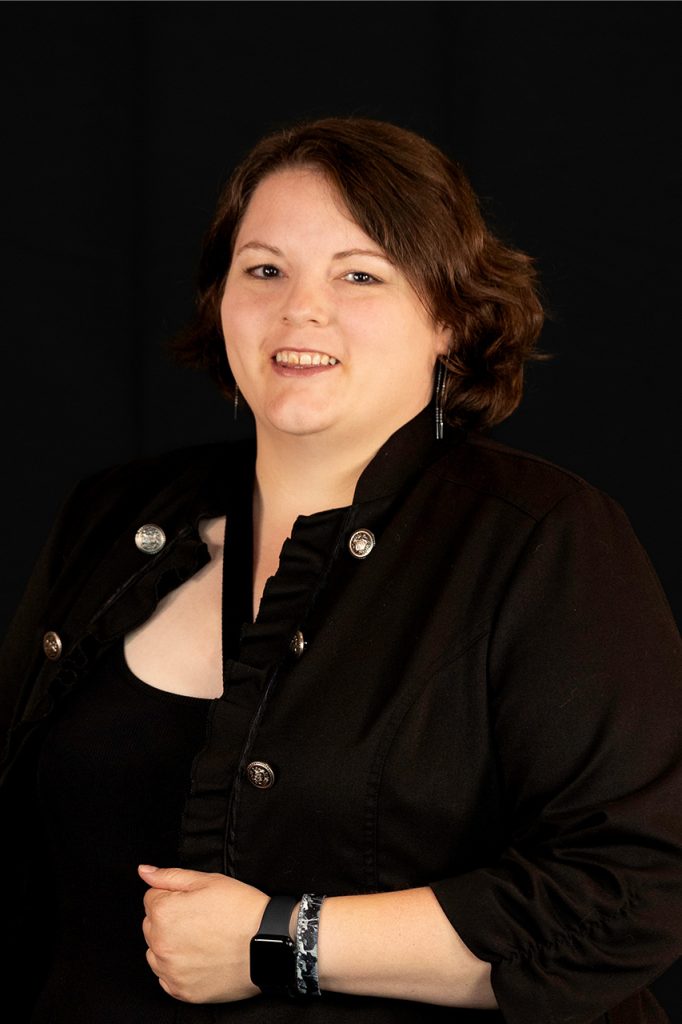 Smiling young adult white woman in a black jacket, posing against a black background.