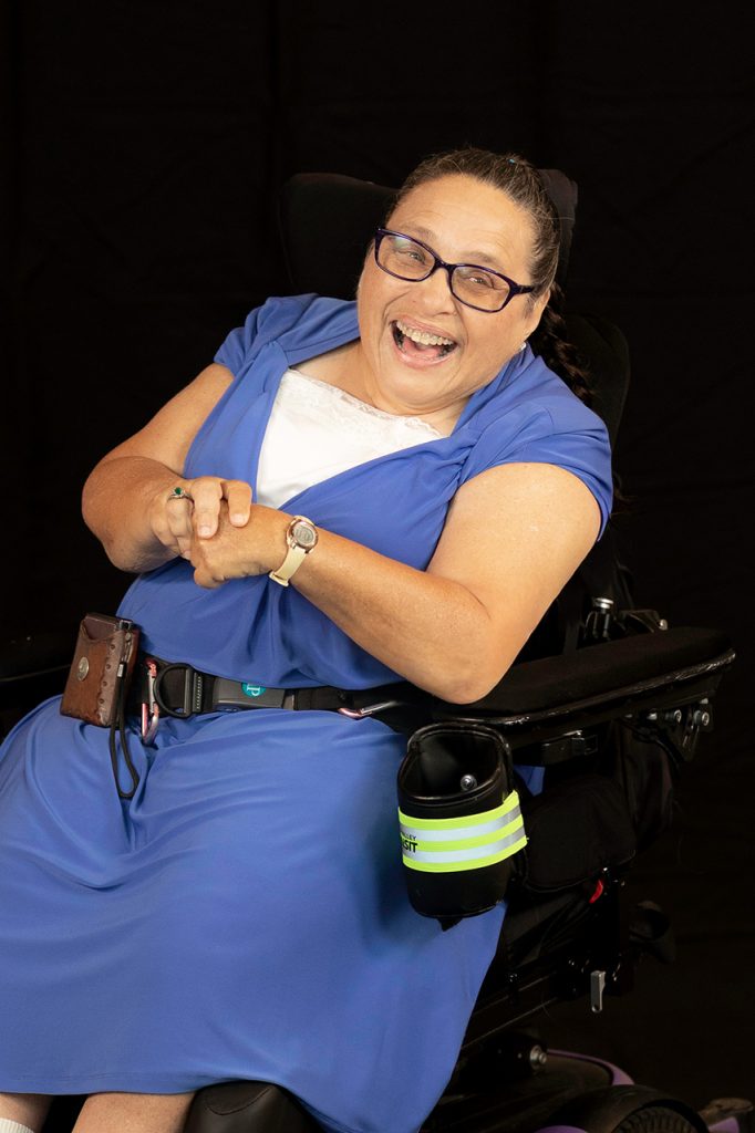 Laughing woman in a wheelchair wearing a blue dress, sitting against a black background.