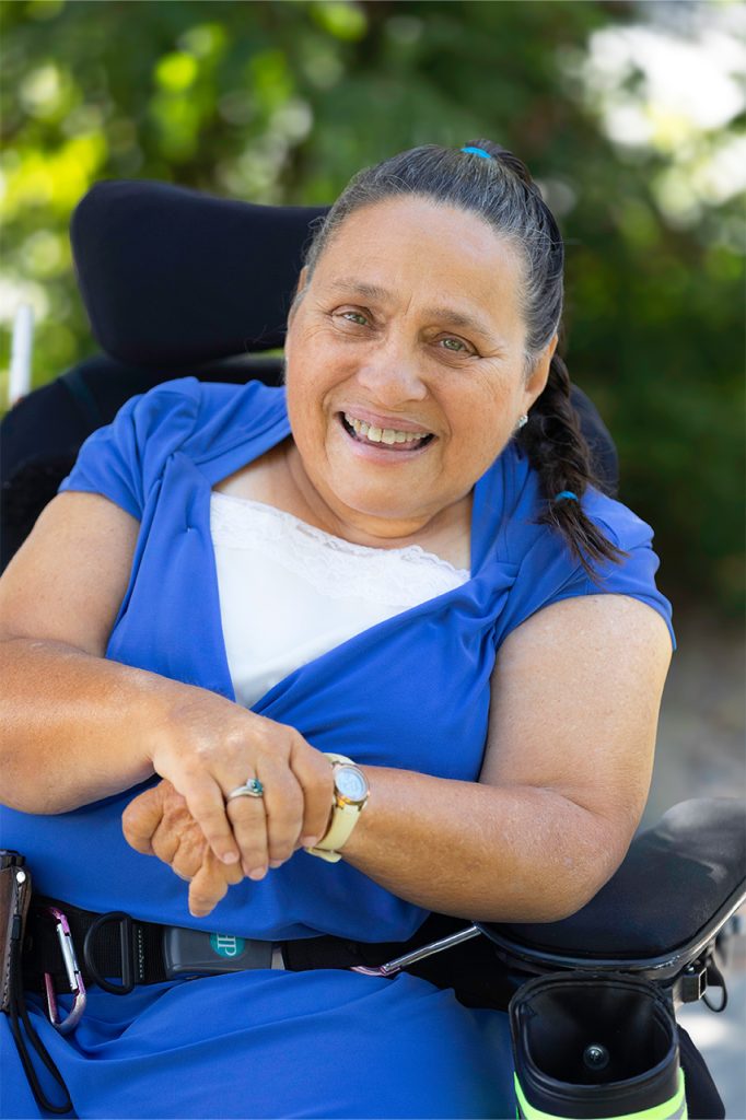 Smiling woman in a wheelchair wearing a blue dress outdoors, surrounded by greenery.