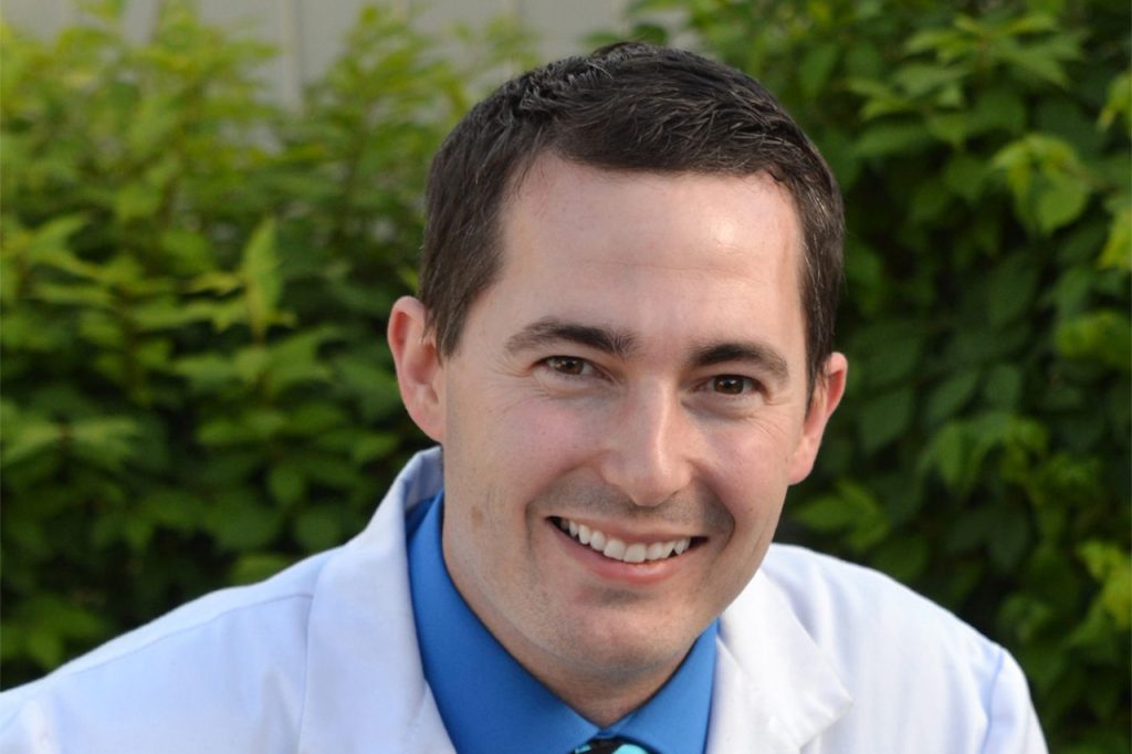 Smiling male health provider in a blue shirt and white coat with green foliage background.