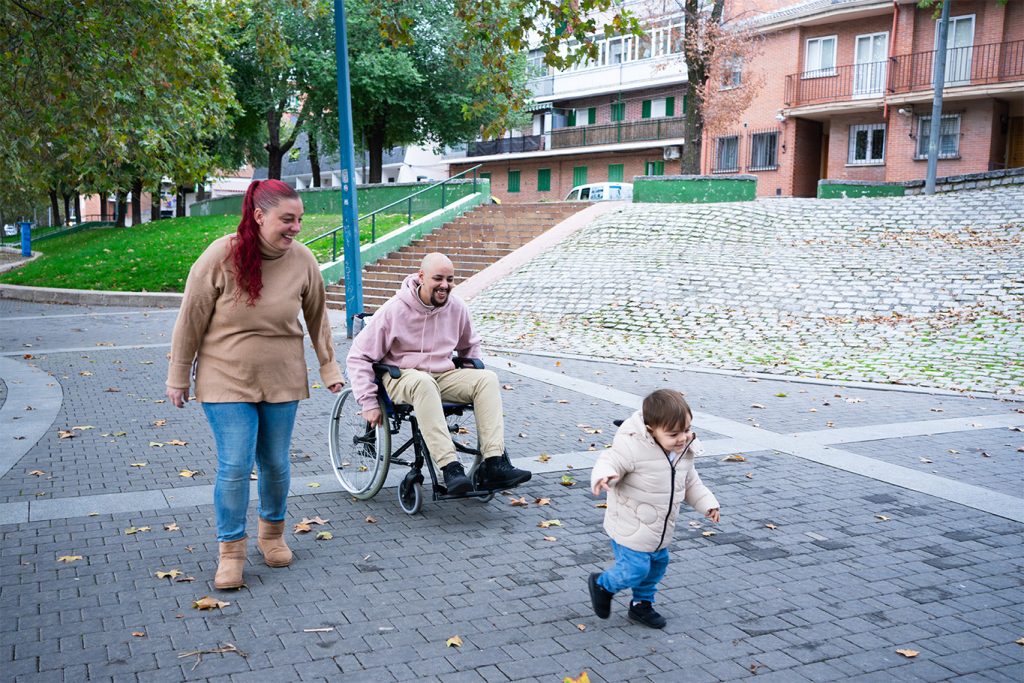 Family enjoying a walk outdoors in an urban residential setting. The child leads, followed by parents, one using a wheelchair.