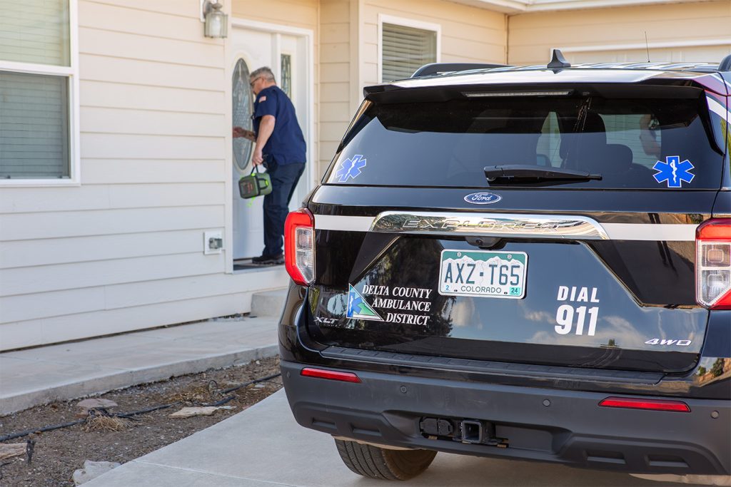 Paramedic entering a patient's home with Delta County Ambulance SUV parked outside. Part of Delta County Ambulance District's mobile integrated health program.